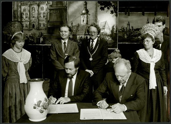 Schwarz-Weiß-Aufnahme: Zwei Männer sitzen an einem Tisch unterzeichnen jeder ein Schreiben. Um sie herum stehen weitere fünf Personen