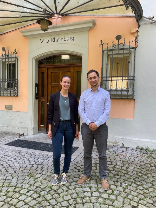 Eine Frau und ein Mann stehen vor dem überdachten Eingang zu einer Villa mit rosa Fassade.