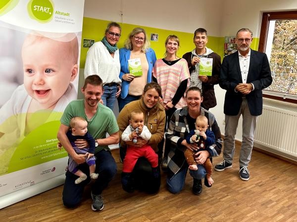 Eine Gruppe Menschen mit drei Babys steht vor einem Plakat. 