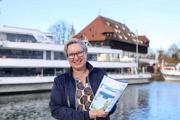 Eine blonde Frau hält ein Dokument in der Hand, im Hintergrund zwei weiße Schiffe.
