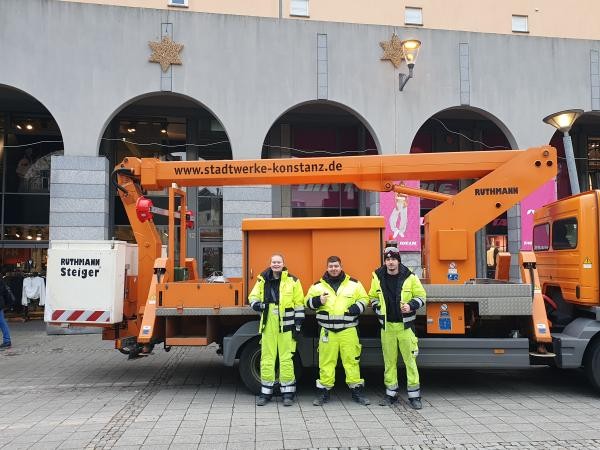 Drei Personen in gelber Arbeitskleidung stehen vor einem orangenem Steiger. 