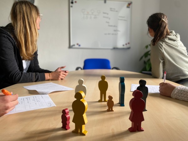 Personen sitzend an einem Tisch, in der Mitte bunte menschliche Holzfiguren 