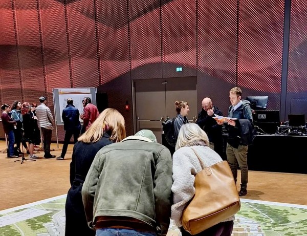 Menschen betrachten großen Plan auf dem Fußboden. Im Hintergrund Personen  im Gespräch.