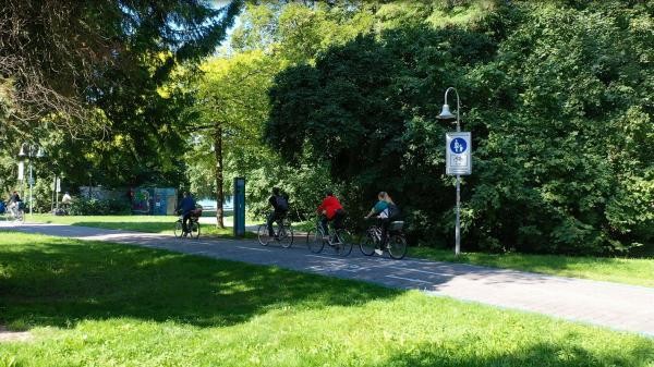 Auf dem Bild ist eine grüne Wiese zu sehen, außerdem 4 Fahrradfahrer, die auf Richtung Westen fahren. Hinter den Radfahrern kann man viele grüne Bäume sehen.