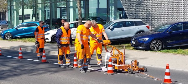 Auf einer großen Straße sind Mitarbeitende der Technischen Betriebe Konstanz in neonorangener Arbeitskleidung im Trupp unterwegs. Mithilfe einer großen, handgeführten orangenen Maschine bringen sie weiße Fahrbahnmarkierungen an.