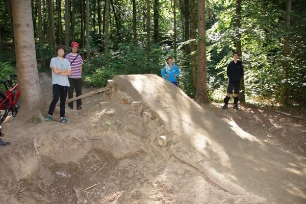 Das Bild zeigt einen Ausschnitt im Wald, eine Schanze für Mountainbikes. Zu sehen sind außerdem noch mehrere Personen.