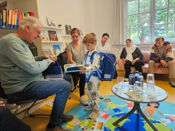 Das Bild zeigt eine Gruppe von Erwachsenen und einem Kind in einem freundlich eingerichteten Raum mit Büchern und Spielsachen. Ein Mann überreicht dem Kind ein Stiftemäppchen und einen Turnbeutel, während andere Personen aufmerksam zusehen. Die Atmosphäre wirkt herzlich und unterstützend.