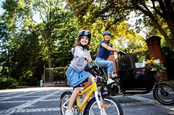Eine Frau auf einem Lastenrad und ein Mädchen auf einem Kinderfahrrad. Im Hintergrund Bäume und der Zaun eines Parks.