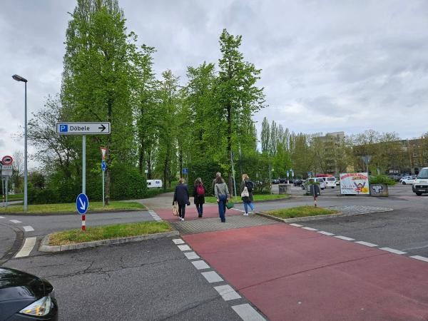 Mehrere Fußgänger überqueren eine Straße mit markiertem Fuß- und Radweg bei einem Kreisverkehr. Ein Wegweiser zeigt die Richtung zum Parkplatz Döbele an. Die Querung ist barrierearm gestaltet mit abgesenktem Bordstein und guter Sichtbarkeit.