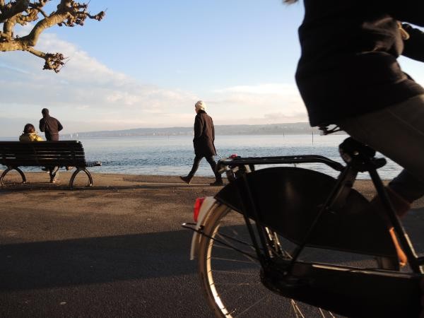 Das Bild zeigt eine Promenade am Wasser bei sonnigem Wetter mit Spaziergängern, einer sitzenden Person auf einer Bank und einem vorbeifahrenden Fahrrad im Vordergrund. Im Hintergrund ist ein ruhiger See mit Blick auf das gegenüberliegende Ufer zu sehen. Die Szene vermittelt eine entspannte Atmosphäre am Seeufer.