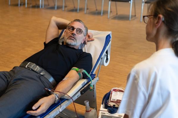 Auf dem Foto liegt ein Mann auf einer Liege. Er spendet Blut. Sein Arm ist ausgestreckt. An seinem Arm ist eine Nadel. Daran hängt ein Beutel mit Blut. Rechts vorne sitzt eine Frau. Man sieht sie von hinten.