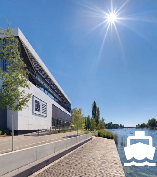 Das Bild zeigt das moderne Konferenzgebäude „Bodenseeforum Konstanz“ bei strahlendem Sonnenschein. Es liegt direkt am Ufer des Rheins, umgeben von Bäumen und einer Promenade. Rechts unten ist ein weißes Symbol für einen Wasserbus eingeblendet.